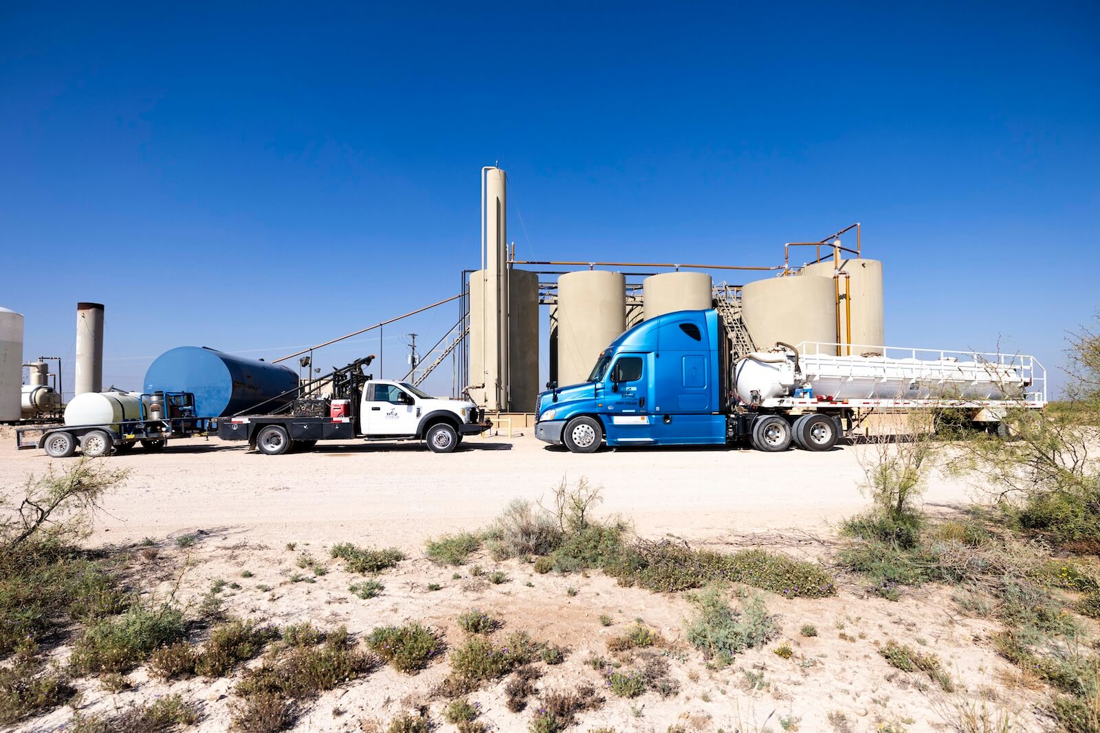 A large blue B-z Construction semi-truck with a tank trailer parked next to a white service truck with a smaller tank trailer at an Oil and gas site.