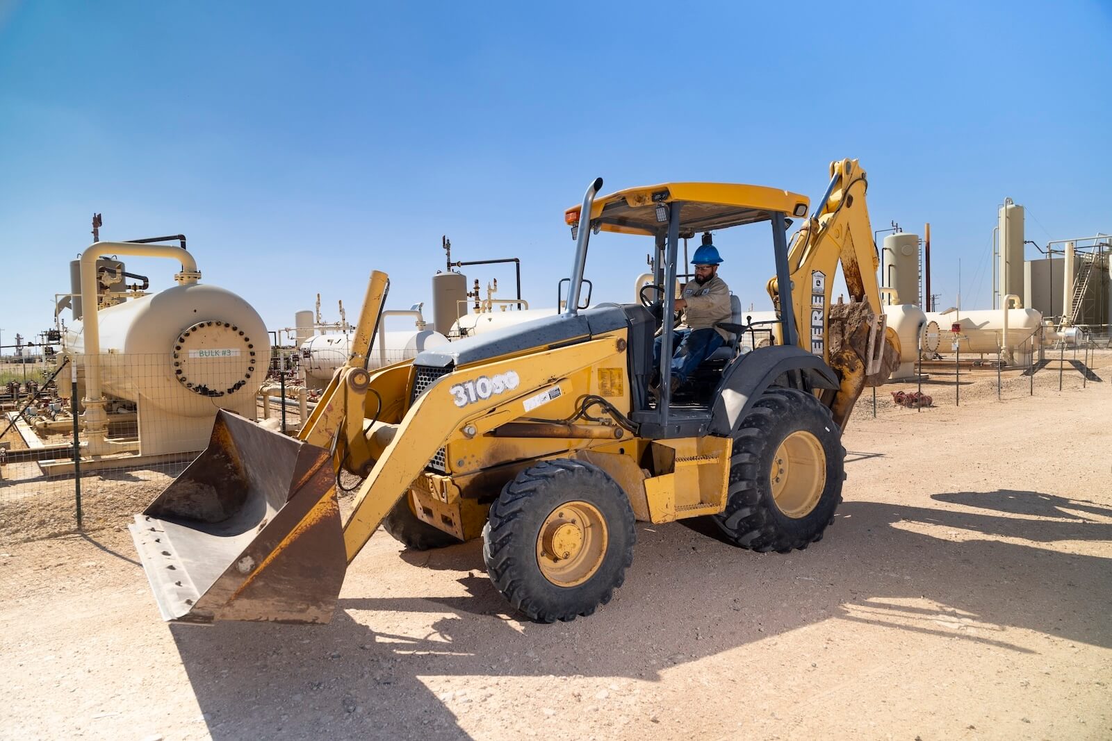 Worker operating a yellow John Deere backhoe loader at West Texas oilfield.