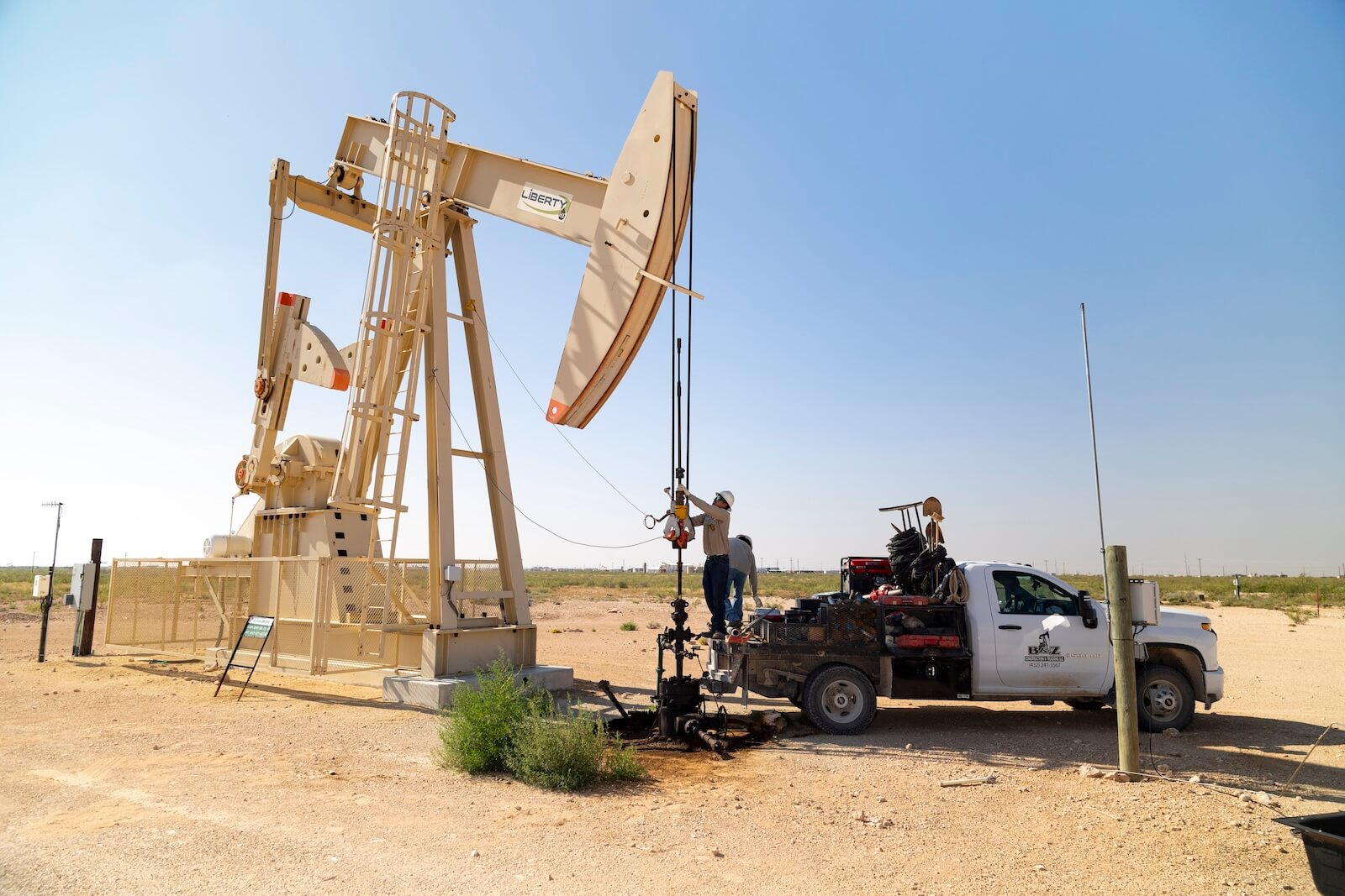 A worker on the back of a B&Z company truck servicing a large oil pumpjack in an open, sunny field.