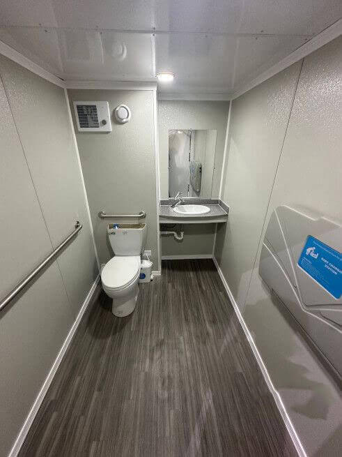 ADA compliant restroom stall interior in a mobile trailer, featuring a toilet, safety grab bars, a single vanity sink, and a wall-mounted baby changing station.