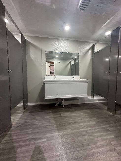 rWide interior view of a portable restroom trailer showing a double-sink vanity, large mirror, and open entrances to the private toilet stalls on either side.