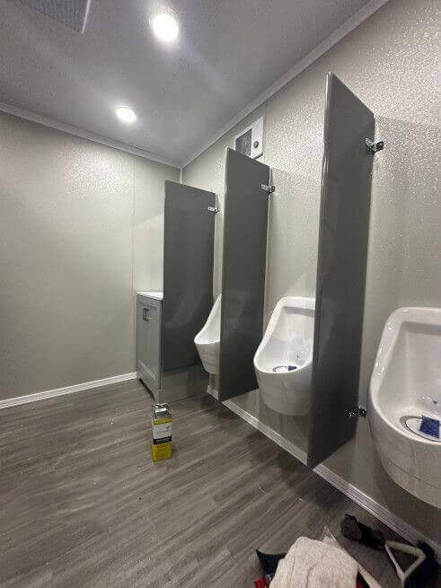 Interior of the men's side of a portable restroom trailer showing three white urinals separated by grey privacy dividers.